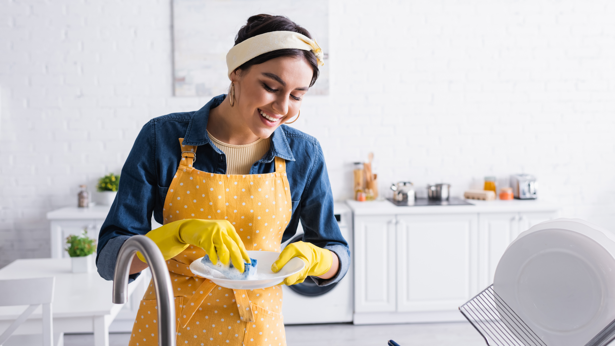 A woman wearing a yellow apron and rubber gloves is washing a plate in a bright, modern kitchen, smiling as she cleans.